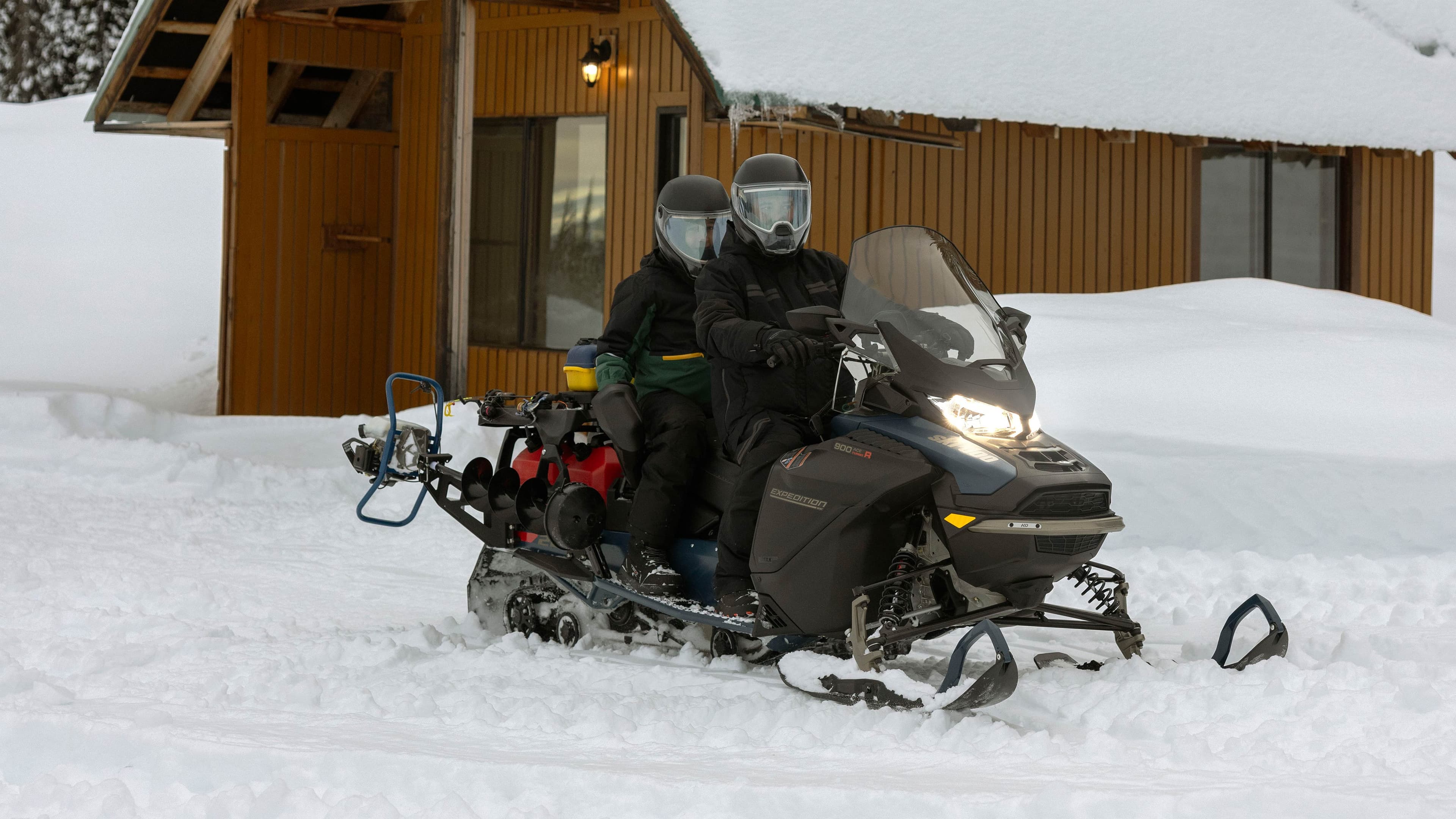 Два человека на Ski-Doo Expedition SE 2025 у деревянного домика зимой